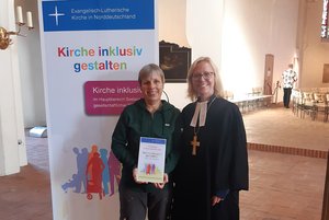 Zwei Frauen vor einem Aufsteller, eine hält eine Urkunde in der Hand